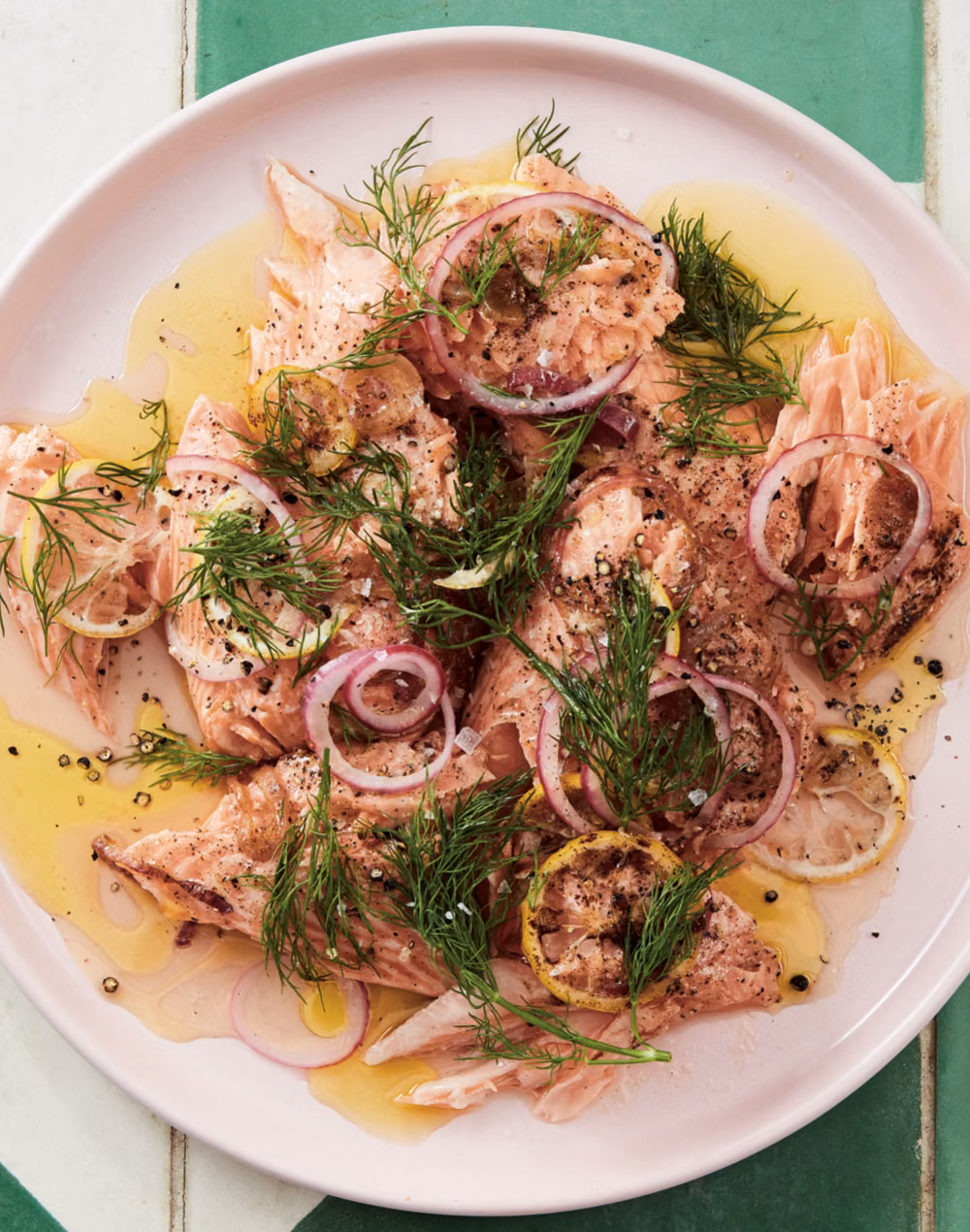 Cooked salmon pieces topped with fresh dill, sliced red onions, lemon, black pepper, and drizzled olive oil on a light pink plate.