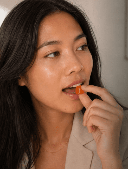 A woman holding a small, orange gummy supplement to her lips.