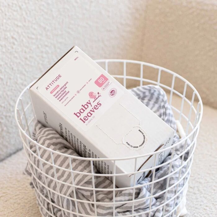 A white box of Attitude Baby Leaves wipes sits in a white wire basket with folded striped and solid cloths underneath.