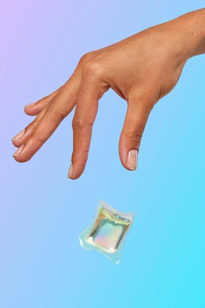 A hand with manicured nails drops a single laundry detergent pod against a blue and purple gradient background.