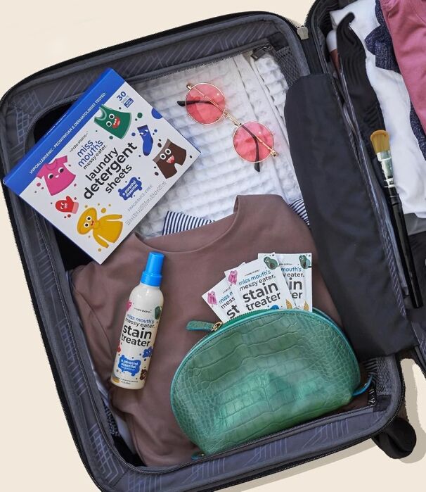 Open suitcase with folded clothes, a green toiletry bag, travel-size detergent sheets, stain treater spray, sunglasses, and makeup brush.