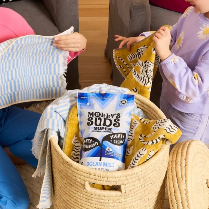 Two people folding towels next to a laundry basket containing a bag of Molly’s Suds Super laundry powder in Ocean Mist scent.