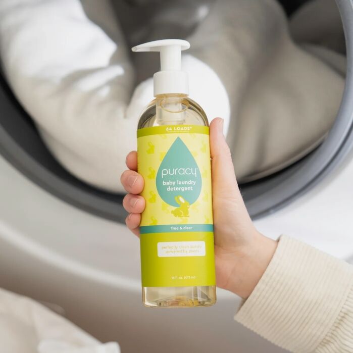 A hand holds a bottle of Puracy baby laundry detergent near the open door of a washing machine with clothing inside.