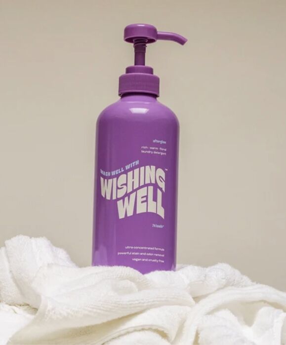 A purple pump bottle labeled “Wishing Well” sits on white towels against a neutral background.
