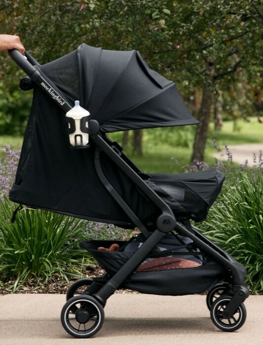 A person pushes a black stroller with a bottle in a holder, along a path bordered by greenery and purple flowers.