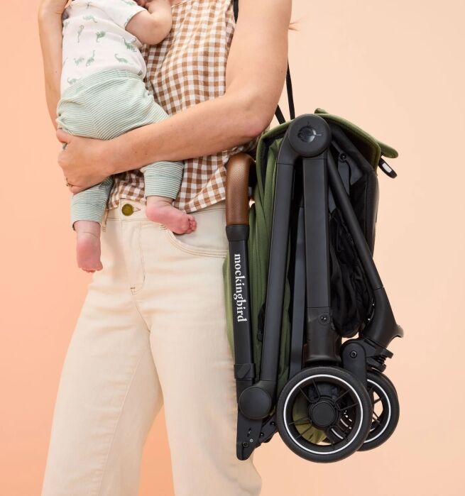 Person holding a baby in one arm and carrying a folded Mockingbird stroller with a shoulder strap against a peach-colored background.