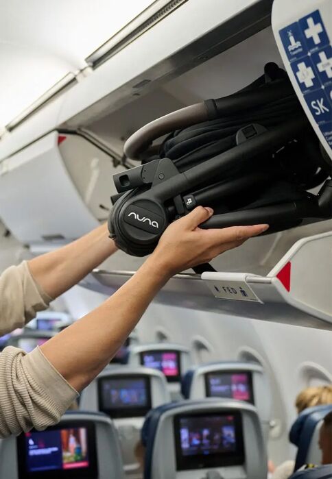 Person placing a folded Nuna stroller into an overhead compartment on an airplane, with passengers seated and screens visible in the background.