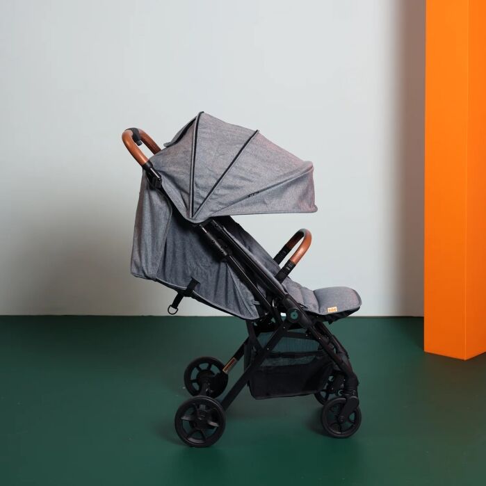 A gray baby stroller with a canopy is positioned sideways on a green floor, next to a tall orange rectangular structure, against a plain white wall.