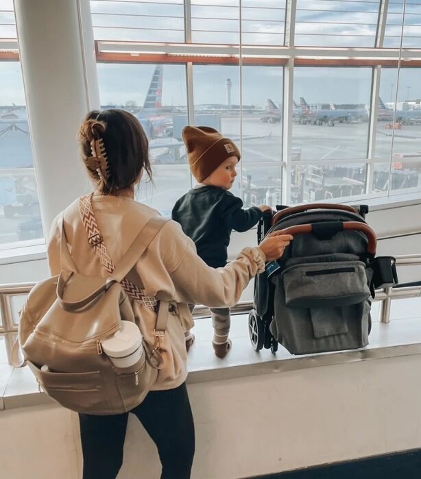A woman with a backpack stands next to a toddler and a stroller at an airport terminal, looking out at airplanes on the tarmac through large windows.