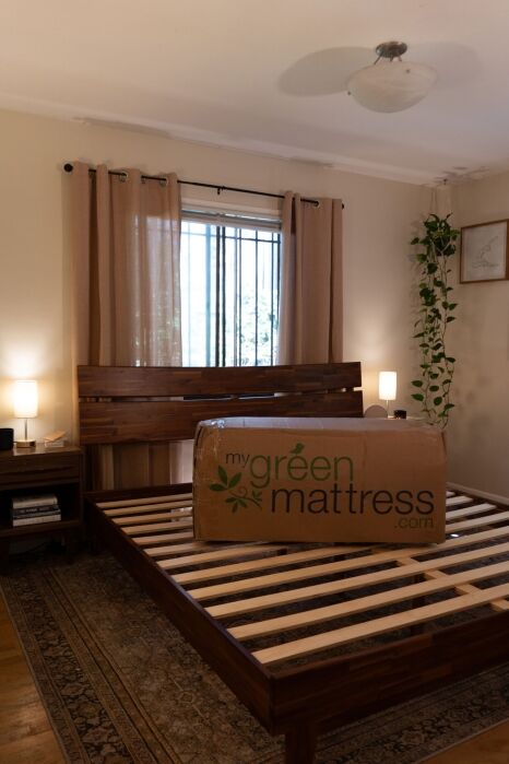 A large cardboard box labeled “My Green Mattress” sits on a wooden bed frame in a neatly arranged bedroom with beige curtains and a potted plant.