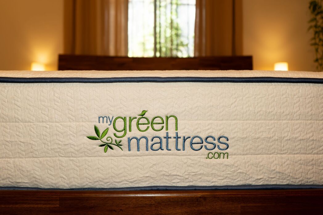 A close-up of a mattress with the logo “mygreenmattress.com” embroidered on the side, set in a softly lit bedroom with a window in the background.