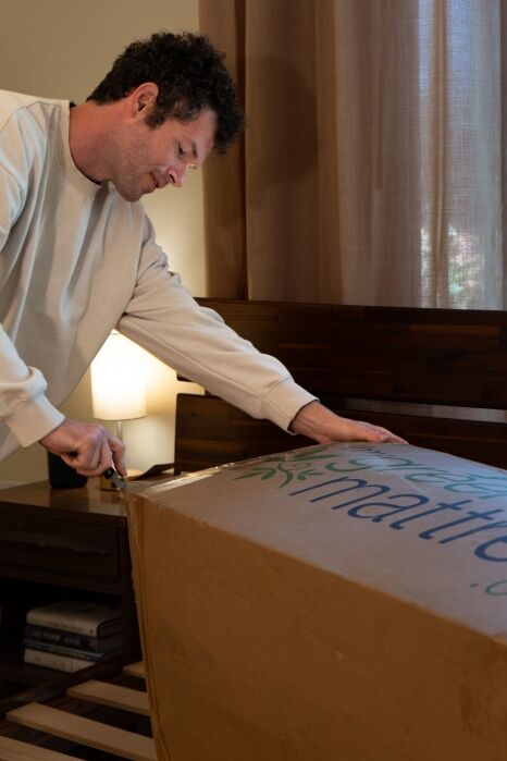 A person uses scissors to open a large cardboard box on a bed frame in a bedroom with a lamp and nightstand nearby.