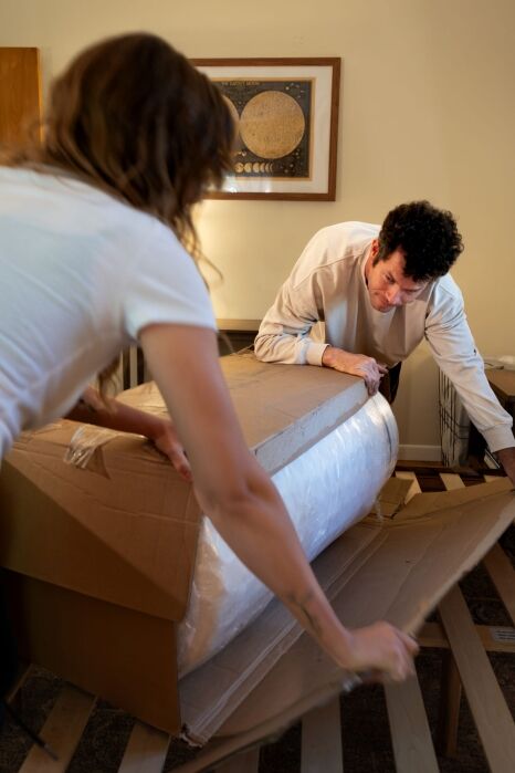 Two people open a large box containing a rolled-up mattress in a bedroom.