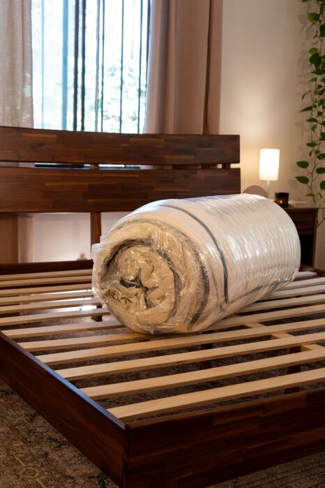 A rolled mattress wrapped in plastic is placed on a wooden bed frame with slats in a bedroom with curtains and indoor plants.