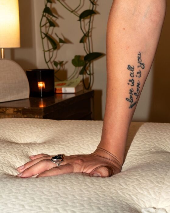 A hand with a large ring presses into a mattress; a forearm tattoo reads “love is all love is you.” A candle, plant, and lamp sit on a bedside table in the background.
