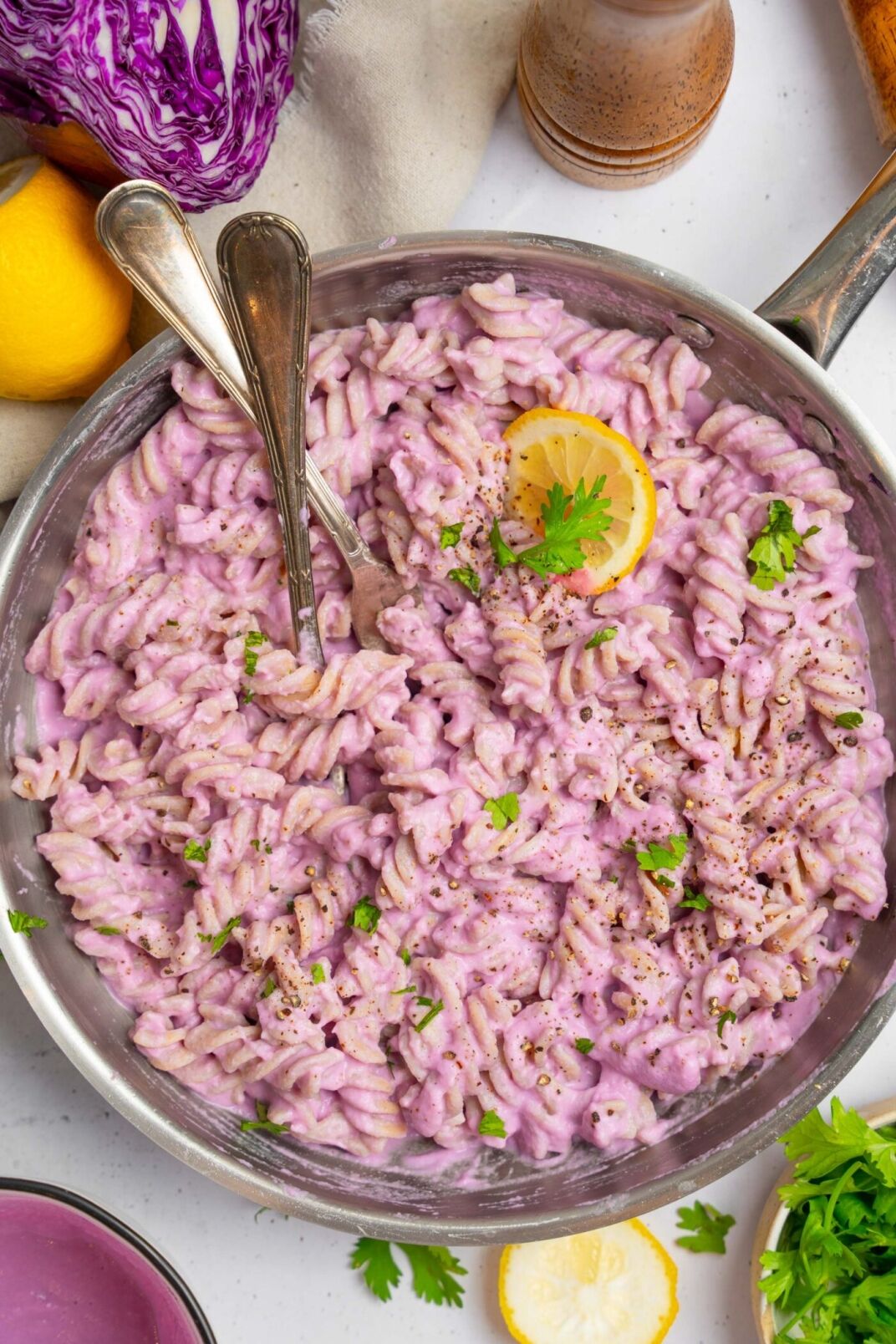 A skillet of rotini pasta in a creamy purple sauce, garnished with parsley, black pepper, and a lemon slice, with two utensils resting in the pan.
