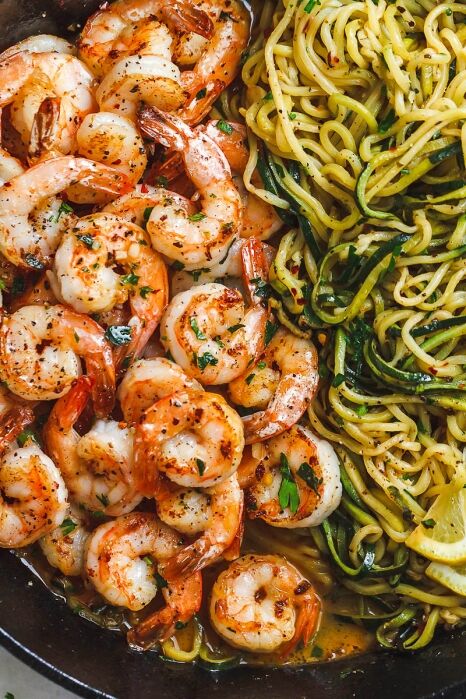 Cooked shrimp seasoned with herbs and pepper sit next to a serving of zucchini noodles in a skillet, garnished with lemon slices and parsley.