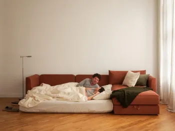A couple lounges together on an unfolded, rust-colored sofa bed in a living room.