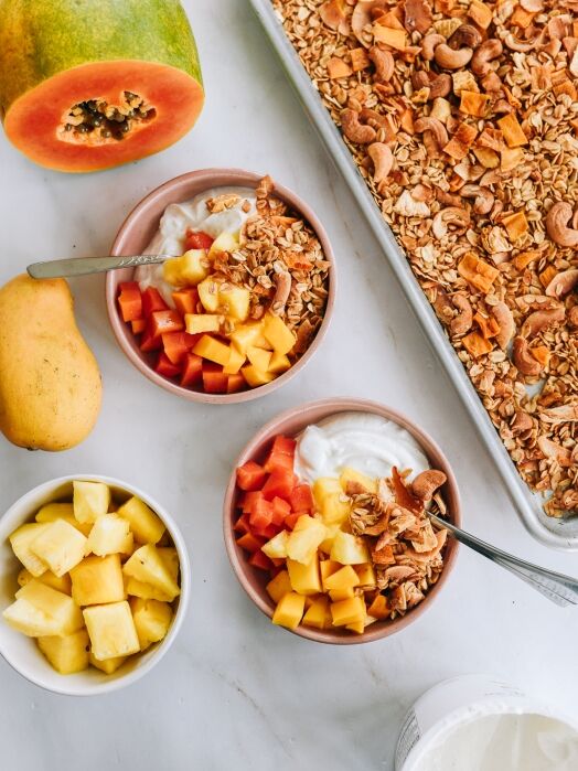 Two bowls of yogurt topped with diced papaya, mango, pineapple, granola, and coconut sit near fresh fruit, a tray of granola, and a container of yogurt on a white surface.