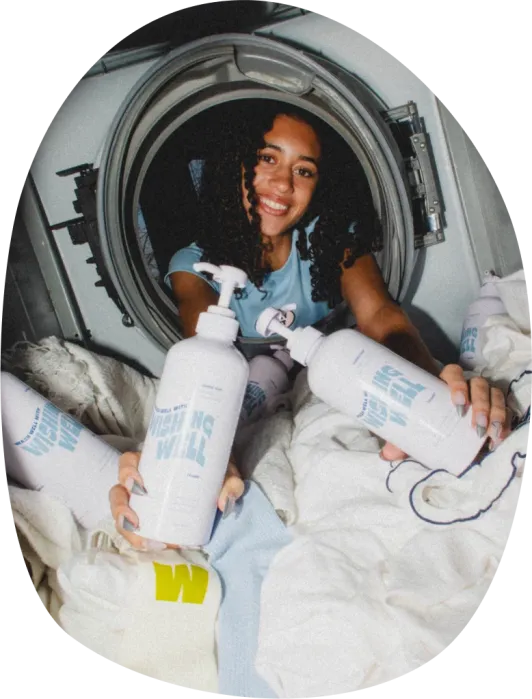 A person sits in a laundry machine surrounded by clothes, holding bottles of laundry detergent and smiling at the camera.