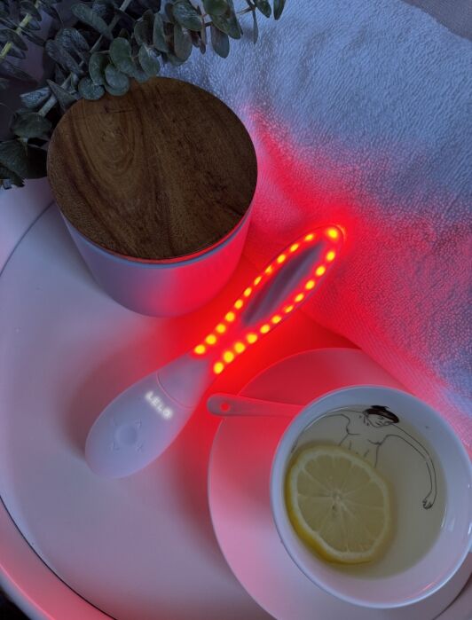 A white LELO LED light therapy wand glowing red on a table next to a cup of lemon water and a lidded container.