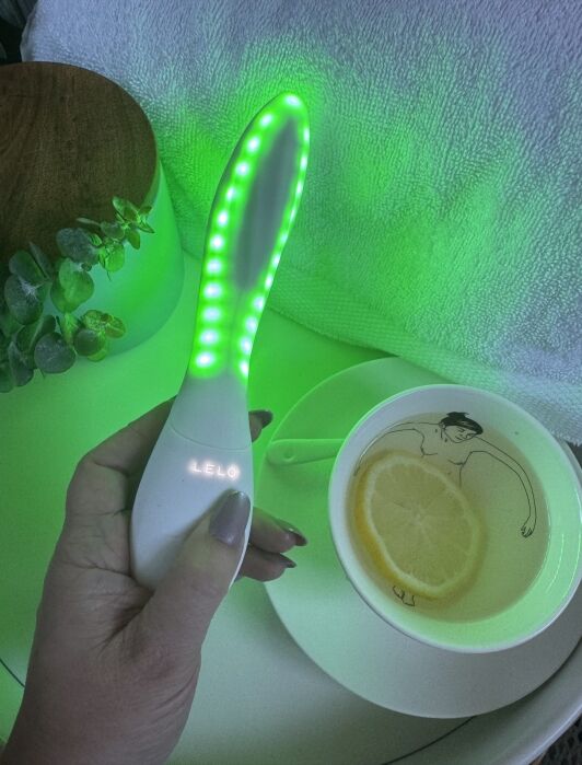 A hand holding a glowing green LELO skincare device next to a teacup with a lemon slice.