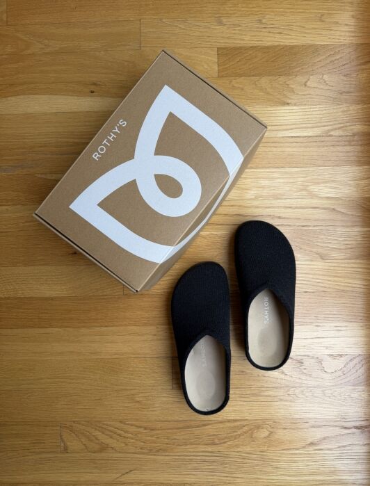 A pair of black Rothy’s slip-on shoes next to their branded cardboard box on a wood floor.