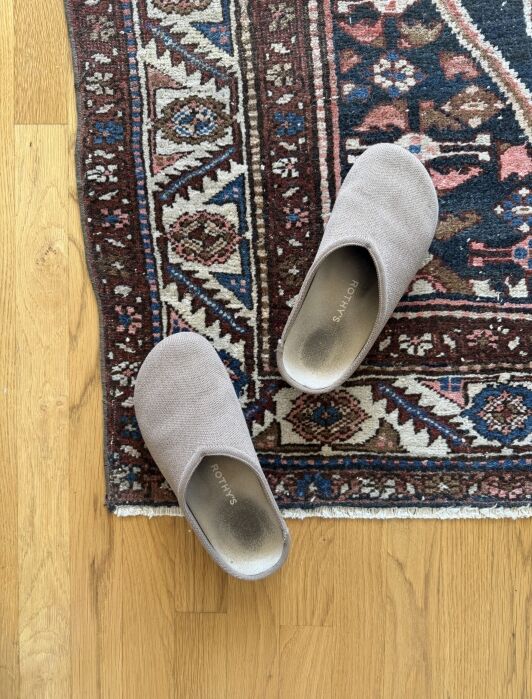 A pair of tan Rothy’s slip-on shoes on a patterned area rug over a wood floor.