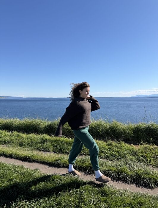 A woman walking along a grassy coastal path with the ocean in the background.
