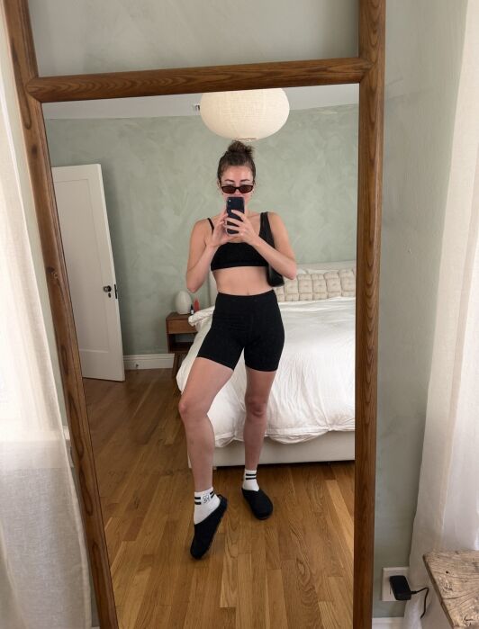 A person in a black sports bra and bike shorts, wearing sunglasses, takes a mirror selfie in a bedroom.