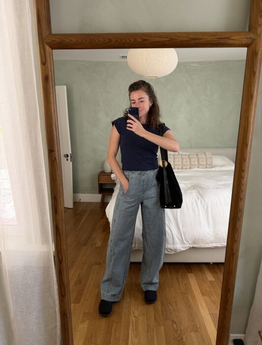 A person takes a mirror selfie wearing a dark blue t-shirt, wide-leg jeans, and a shoulder bag.
