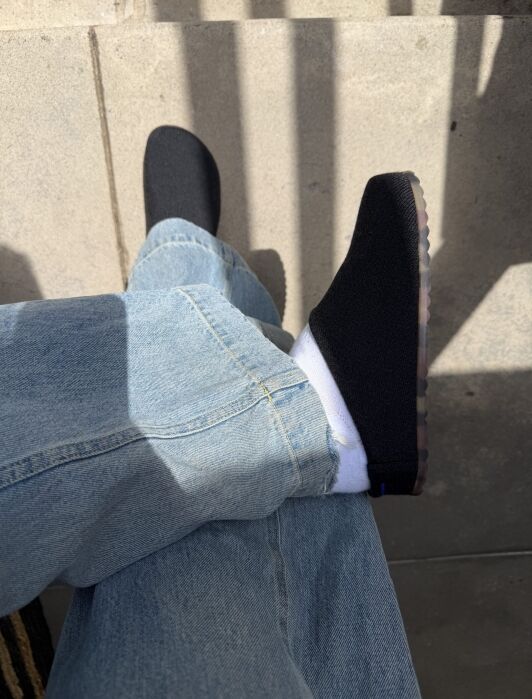 A person wearing light blue jeans, white socks, and black slip-on shoes with translucent soles.