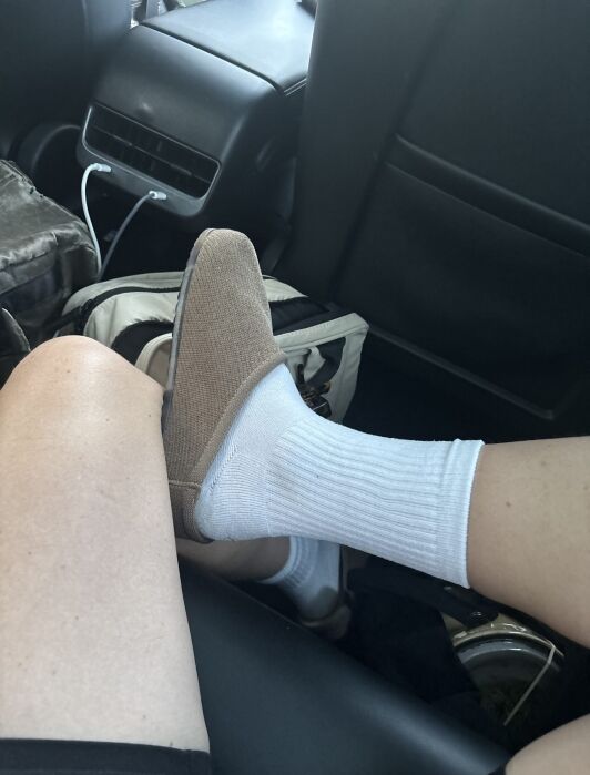 A person’s foot wearing a white sock and a tan slip-on shoe inside a car.