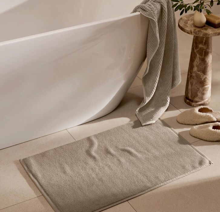 A beige bath mat on tiled floor beside a white bathtub, with a towel draped over the tub and slippers and a small round table nearby.