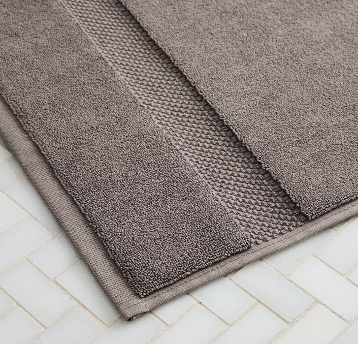 Close-up of a folded taupe bath towel with a textured border, lying on a white tile floor arranged in a herringbone pattern.