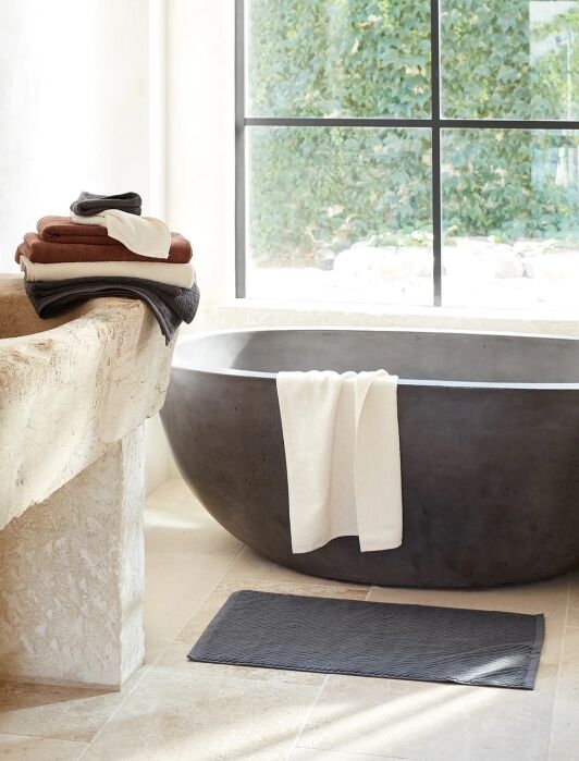 Modern bathroom with a stone tub, folded towels on a stone sink, a white towel draped on the tub, and a dark bath mat on the tiled floor near a large window.