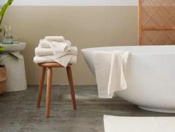 A stack of folded white towels sits on a wooden stool beside a modern white bathtub, with another towel draped over the tub.