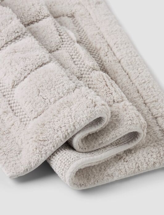 Two light beige, textured bath mats are partially folded on a plain surface. The mats have a plush, patterned surface and appear soft and absorbent.
