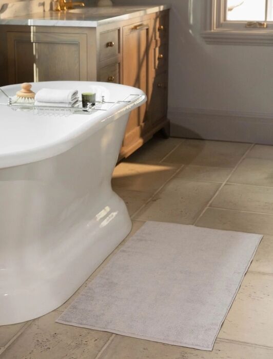 A white bath mat is placed on a tiled bathroom floor next to a white freestanding bathtub, with a tray holding towels and toiletries resting on the tub.