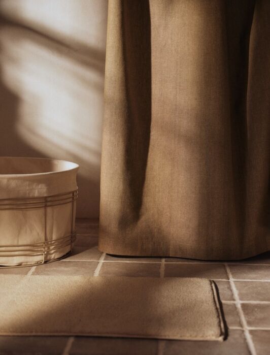 A fabric laundry basket, a hanging curtain, and a rectangular mat are arranged on a tiled floor in soft, natural light.