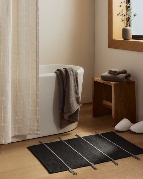 Minimalist bathroom with a white tub, beige shower curtain, dark towels, a wooden stool, folded towels, a vase with branches, white slippers, and a black striped rug on a wooden floor.