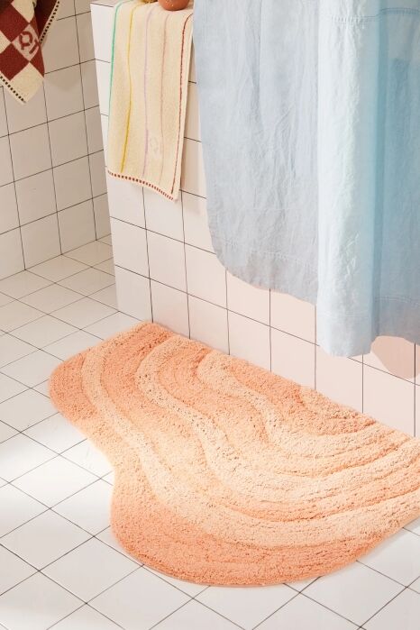 Peach-colored wavy bath mat on white tiled floor next to a blue shower curtain and hanging towels.