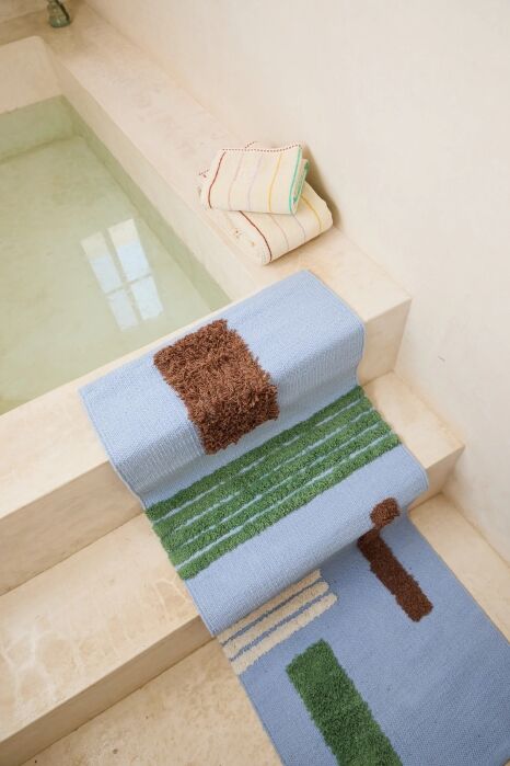 A blue bath mat with green and brown abstract patterns is draped over white stone steps next to a pool; two folded towels rest on the top step.