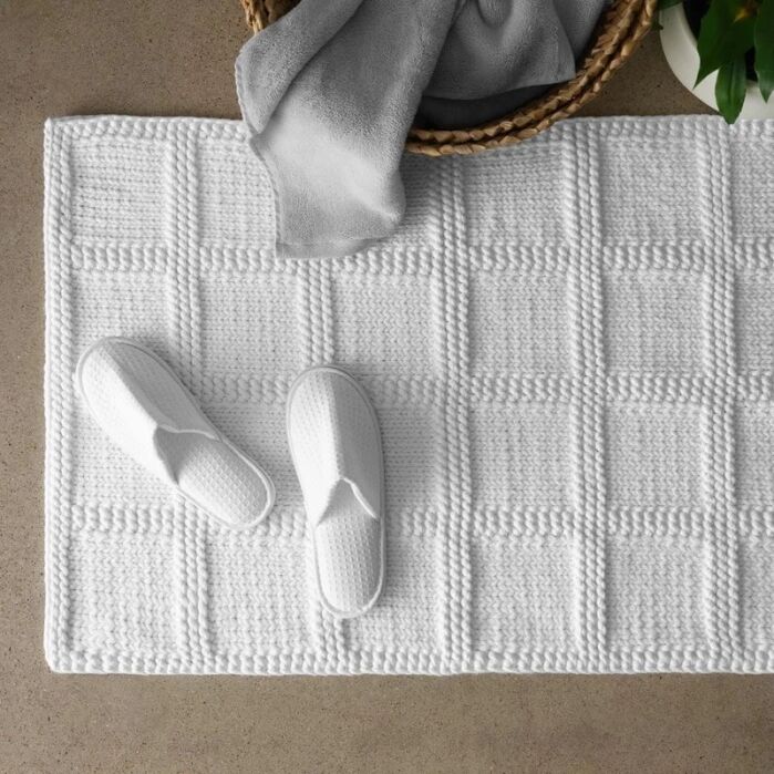 White textured bath mat with a pair of white slippers placed on it, next to a woven basket holding a gray towel; green leaves partially visible.