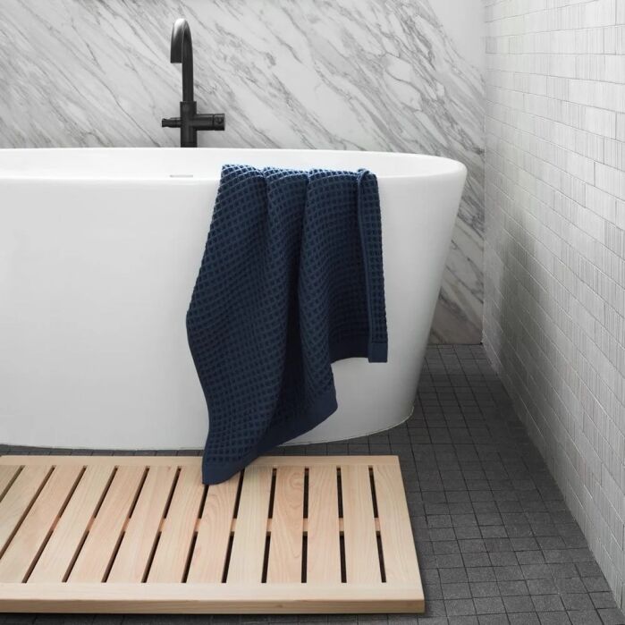 A white bathtub with a black faucet, a dark blue textured towel draped over the edge, and a wooden bath mat on a dark tiled floor. Marble and tile walls are in the background.