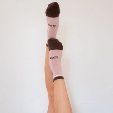 A person’s bare legs are raised against a plain white wall, wearing mismatched pink and brown organic socks labeled “EARTH” and “COZY.”.