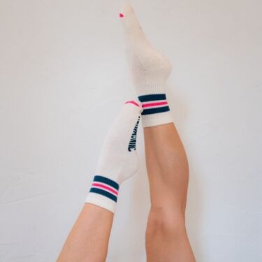 Two legs raised against a white wall, wearing organic socks with blue, red, and pink stripes, and text partially visible on the sides.