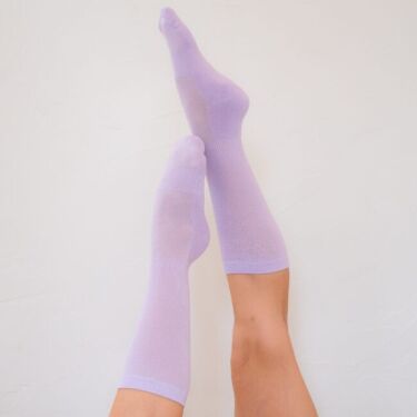 Two legs are raised against a white wall, wearing light purple knee-high organic socks.