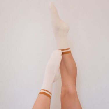 Two legs raised and crossed against a white wall, wearing organic socks with orange stripes and denim shorts.