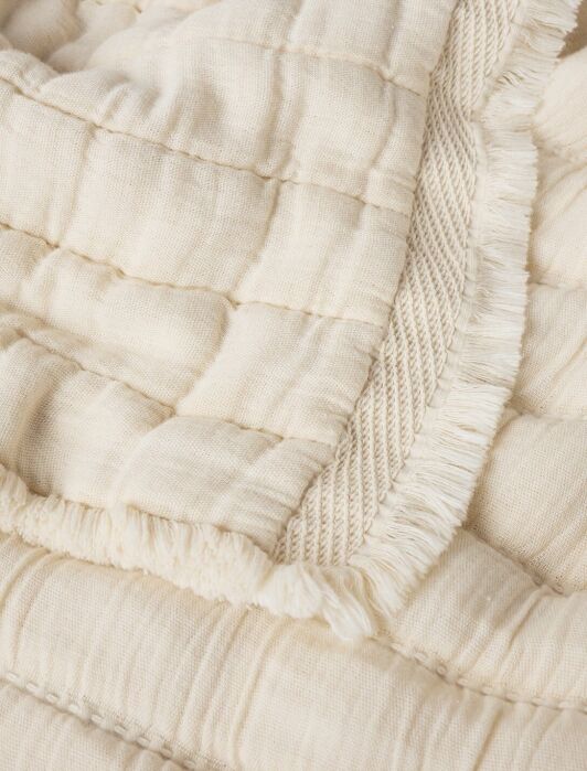 Close-up of a cream-colored, quilted fabric blanket with a textured surface and fringed edge detailing.